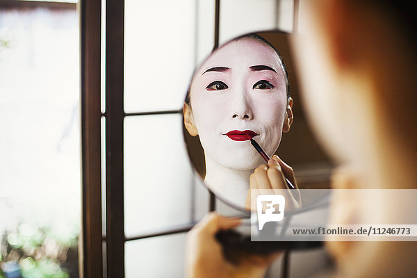 Geisha-Frau mit traditioneller weißer Gesichtsschminke  die mit einem Pinsel und einem Spiegel leuchtend roten Lippenstift aufträgt.