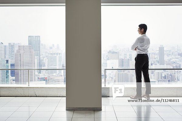 A businessman in the office standing by a large window with arms folded  looking over the city.