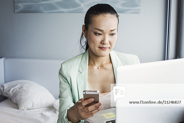 A business woman preparing for work  sitting on a bed using a laptop and holding her smart phone.