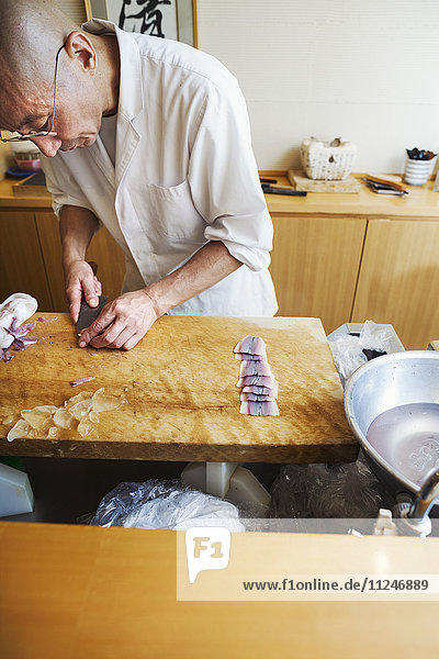 Ein Koch  der in einer kleinen Großküche arbeitet  ein Itamae oder Meisterkoch  der Fisch mit einem großen Messer in Scheiben schneidet  um Sushi zuzubereiten