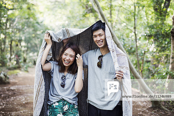 Junge Frau und Mann stehen in einem Wald und halten sich eine Decke über den Kopf.