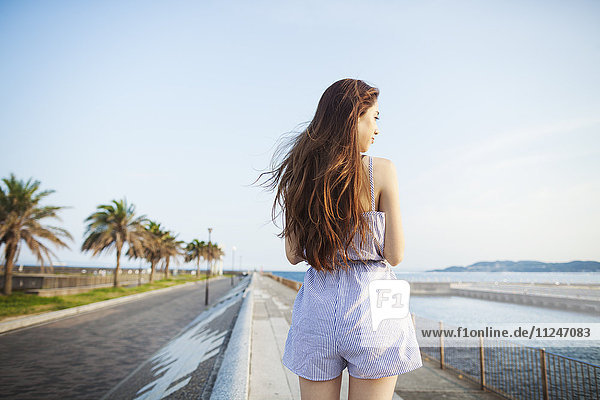 Rückenansicht einer jungen Frau mit langen roten Haaren  die im offenen Raum an einer Straße an der Küste steht.