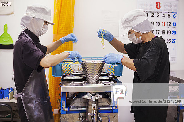 Arbeiterinnen und Arbeiter in Schürzen und Handschuhen beim Wiegen und Verpacken frisch zubereiteter Nudeln in einer Soba-Nudel-Produktionseinheit.