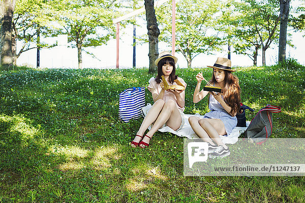 Zwei junge Frauen mit langen braunen Haaren sitzen auf einem Rasen.