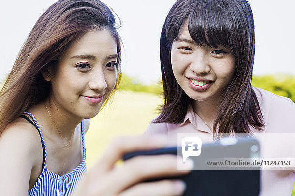 Zwei lächelnde junge Frauen mit langen braunen Haaren  die ein Mobiltelefon in der Hand halten und Selfie nehmen.