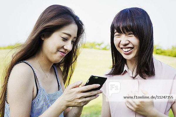 Zwei lächelnde junge Frauen mit langen braunen Haaren  die ein Mobiltelefon in der Hand halten.