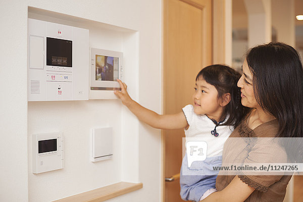 Familienhaus. Eine Frau und ihre Tochter schauen auf einen Bildschirm an der Wand  auf die Bedienelemente eines Haustelefons oder einer Tür.