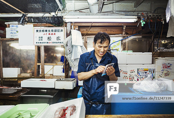 Ein traditioneller Frischfischmarkt in Tokio. Ein Mann in einer blauen Schürze steht hinter der Theke seines Standes und benutzt ein Smartphone  das mit schützendem Plastik überzogen ist.