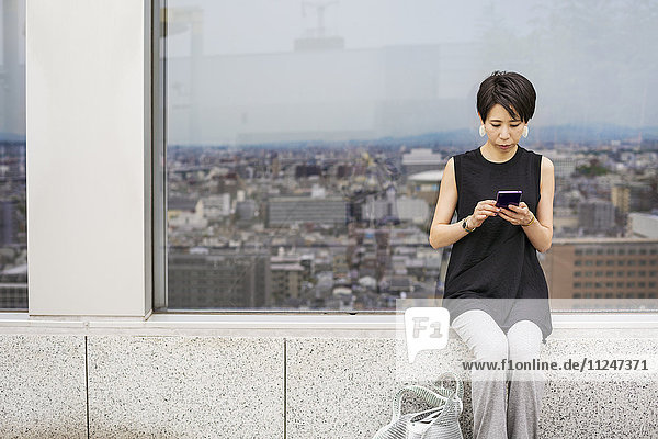 Eine Frau  die an einem Fenster mit Blick auf eine Großstadt sitzt  mit dem Rücken zur Aussicht  mit ihrem Smartphone.
