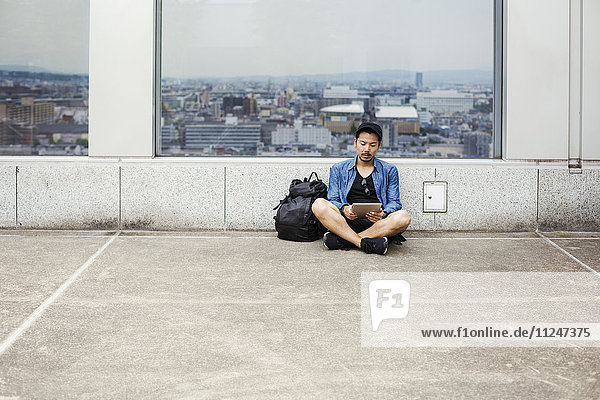 Ein Mann sitzt auf dem Boden und benutzt sein Smartphone vor einem Aussichtsfenster mit Blick auf eine Stadt.