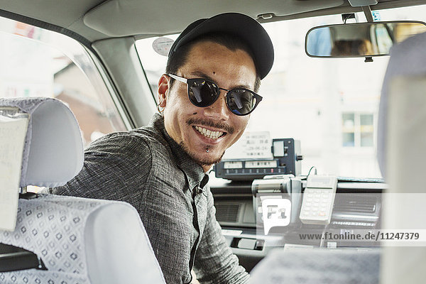 A man wearing sunglasses and baseball hat in the passenger seat of a car turning around and smiling.