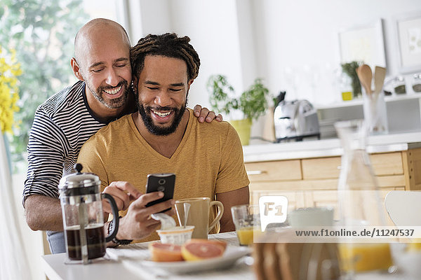 Ein lächelndes homosexuelles Paar benutzt ein Smartphone am Esstisch