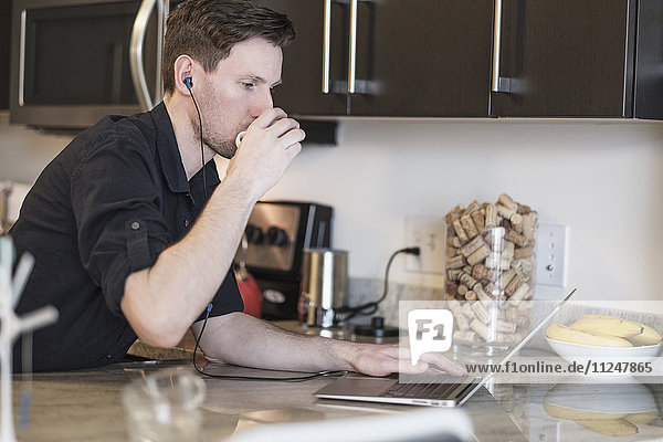 Man drinking while working on laptop computer in kitchen