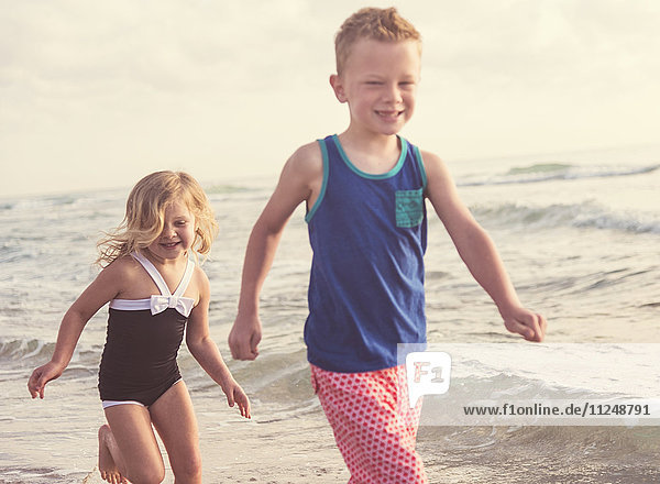 Boy (6-7) and girl (4-5) running on beach by water