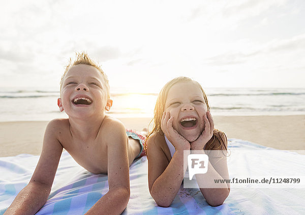 Junge (6-7) und Mädchen (4-5) liegen lachend am Strand