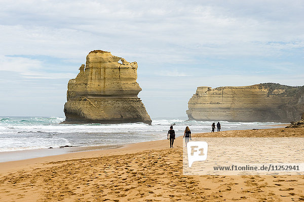 People walk along a beach with one of the Twelve Apostles geological formation in the background  Victoria  Australia  Pacific