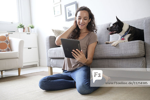 Junge Frau im Wohnzimmer  die ein digitales Tablet mit ihrem Hund benutzt