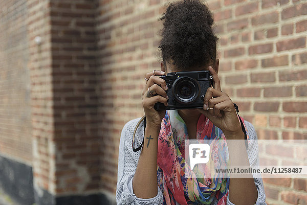 Young woman taking picture with digital camera against brick wall