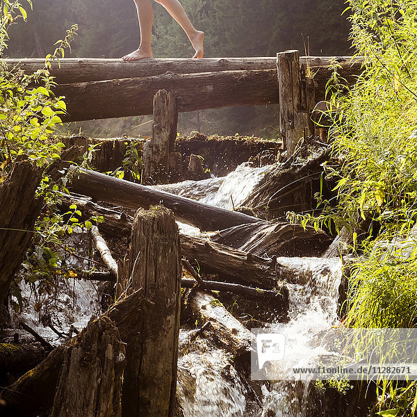 Beine einer kaukasischen Frau  die auf einem Baumstamm geht und einen Wasserfall überquert