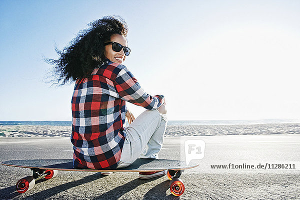 Hispanische Frau sitzt auf einem Skateboard am Strand