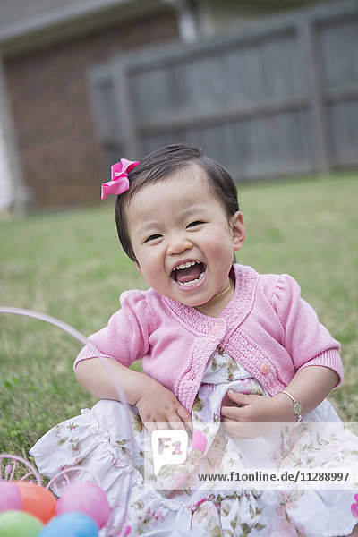 Porträt von Kleinkind Mädchen trägt rosa und sitzt auf Gras lachend mit Osterkorb im Hinterhof