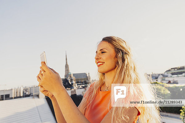 Österreich  Wien  lächelnde  blonde Frau nimmt Selfie auf Dachterrasse mit Stephansdom im Hintergrund