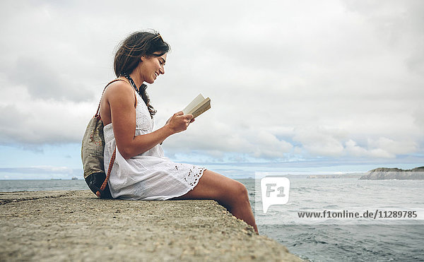 Smiling woman with backpack sitting on pier reading a book