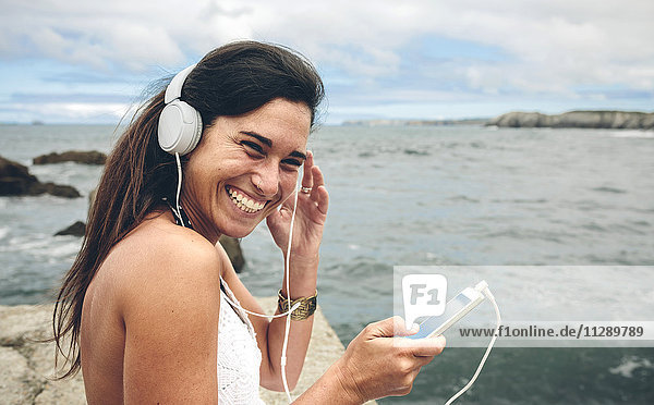 Lachende Frau beim Musikhören mit Kopfhörern vor dem Meer