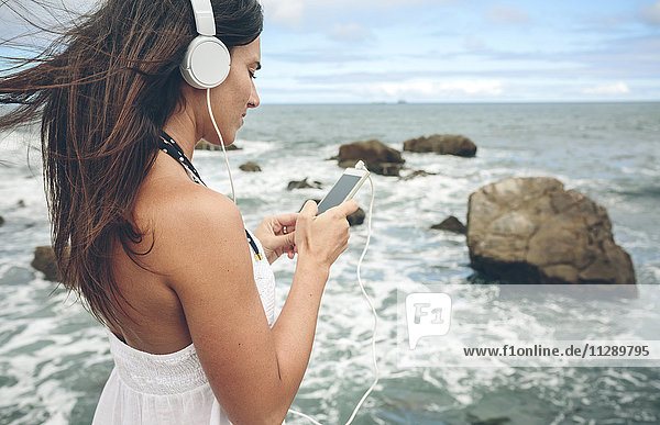 Frau beim Musikhören mit Kopfhörer und Smartphone vor dem Meer