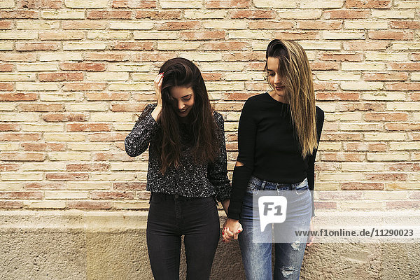 Two women holding hands in front of brick wall looking down