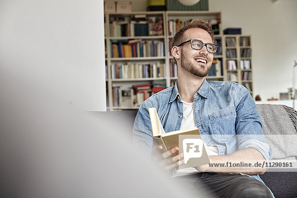 Lachender Mann sitzend mit Buch auf Couch im Wohnzimmer