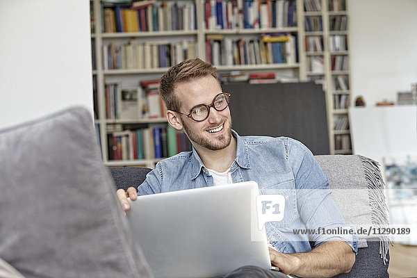 Porträt eines lächelnden Mannes auf der Couch mit Laptop im Wohnzimmer