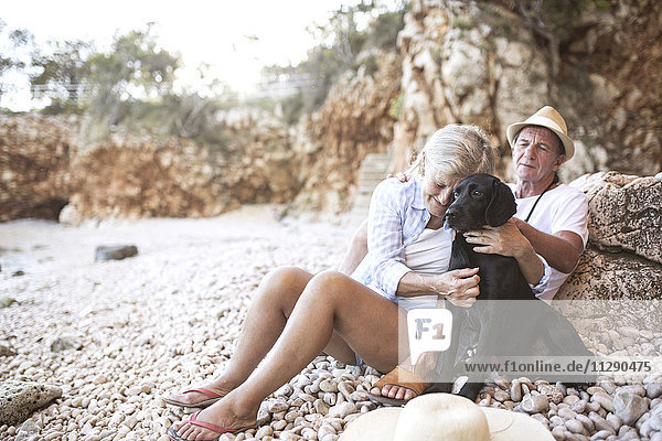 Frau kuschelt Hund am Strand  während ihr Mann sie beobachtet.