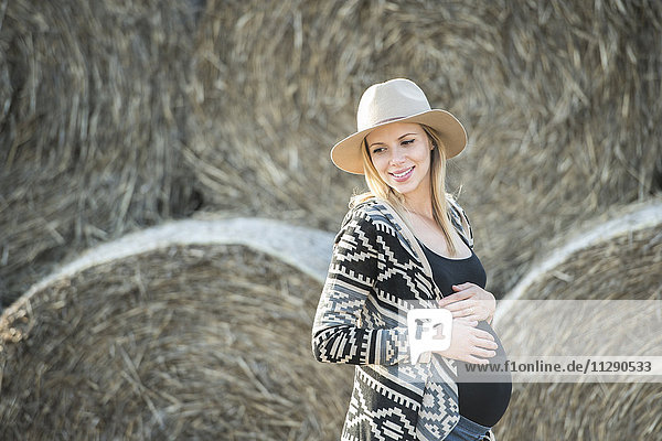 Lächelnde schwangere Frau vor Strohballen stehend