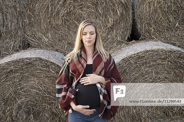 Schwangere Frau mit geschlossenen Augen vor Strohballen stehend