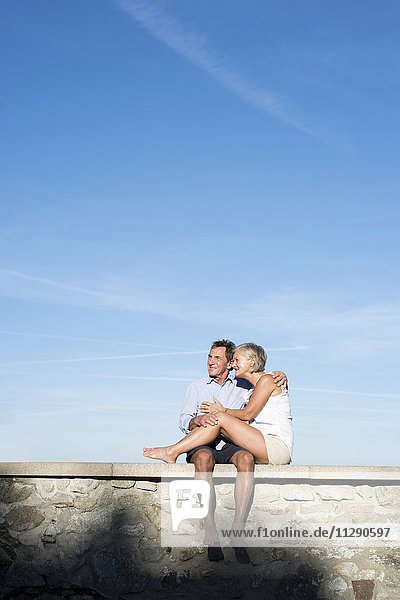 Fröhliches Seniorenpaar sitzt barfuß auf einer Wand vor dem Himmel