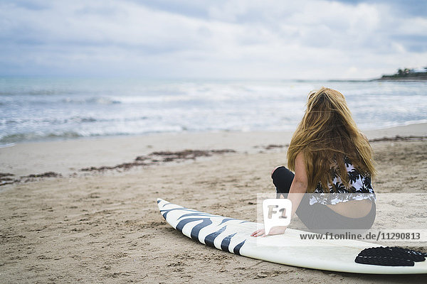 Junge Frau mit Surfbrett am Strand sitzend mit Blick aufs Meer