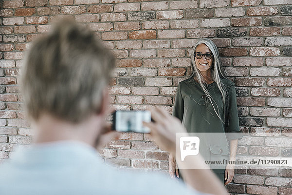 Man taking picture of smiling woman with long grey hair at brick wall