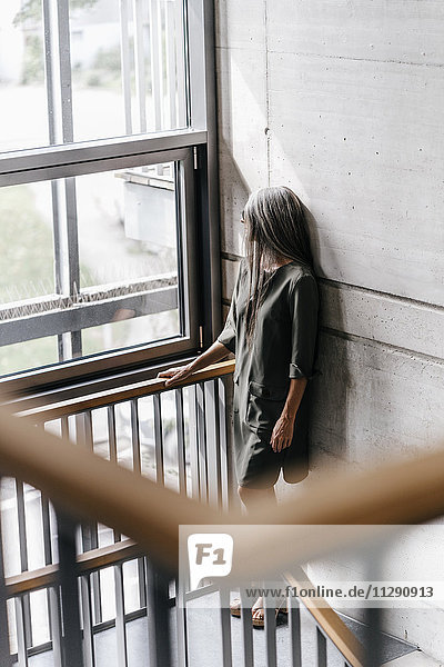Frau mit langen grauen Haaren schaut aus dem Fenster im Treppenhaus