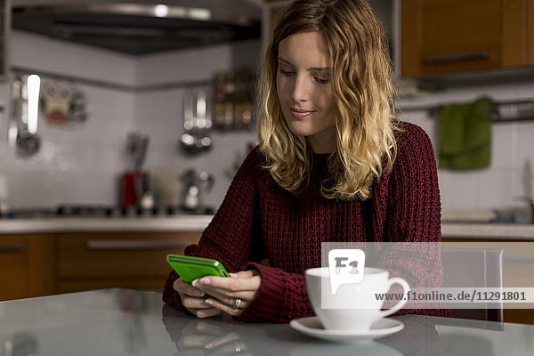 Woman sitting in the kitchen with cup of tea using cell phone