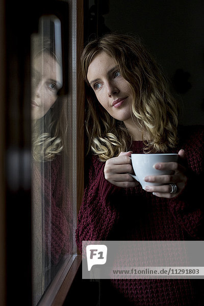 Porträt einer Frau mit Tasse Tee durchs Fenster schauend