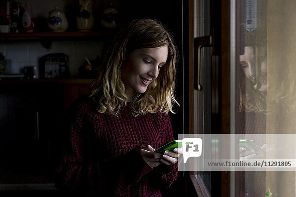 Lächelnde Frau steht zu Hause am Fenster und schaut auf das Smartphone.