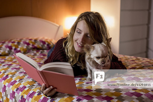 Lächelnde Frau liegt mit ihrem Hund auf dem Bett und liest ein Buch.