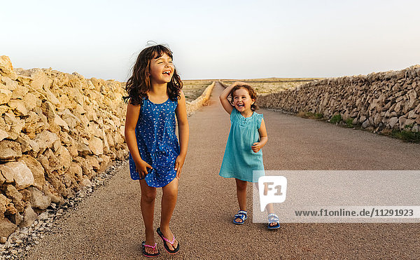 Two laughing little sisters standing on empty road at sunset
