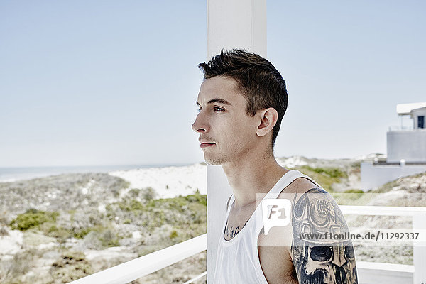 Tätowierter junger Mann auf einer Terrasse des Strandhauses mit Blick auf das Meer