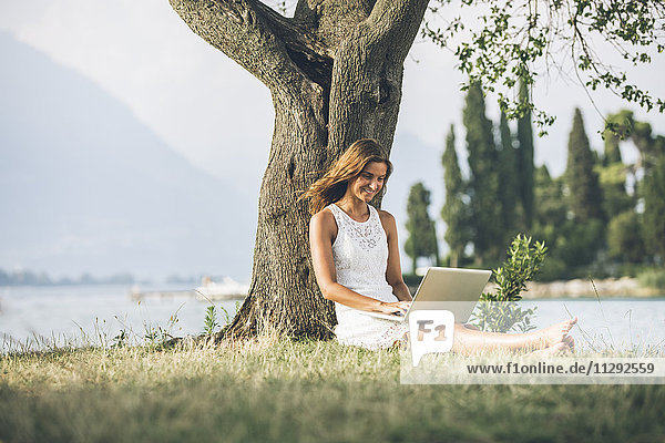 Italien  Gardasee  junge Frau  die sich mit dem Laptop an einen Baum lehnt