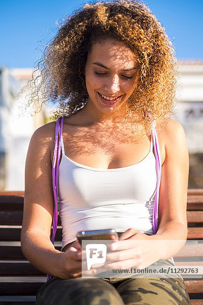 Lächelnde junge Frau sitzt auf der Bank und schaut auf das Smartphone.