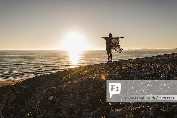 Rückansicht eines fröhlichen Mädchens mit ausgestreckten Armen  das bei Sonnenuntergang auf dem Hügel steht und auf das Meer blickt.