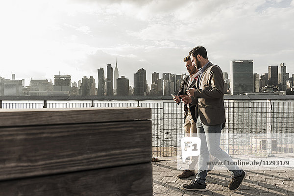 USA  New York City  zwei Geschäftsleute  die am East River spazieren gehen und das Handy anschauen