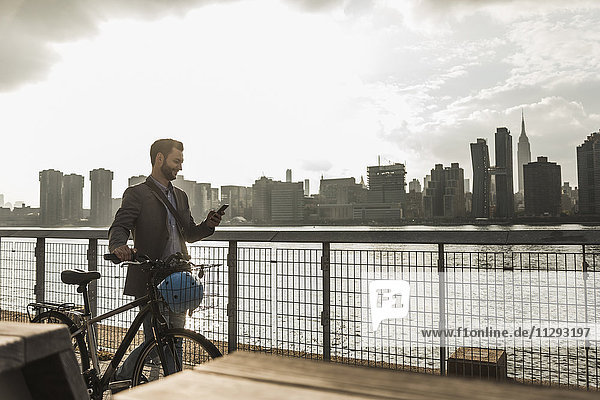 USA  New York City  Geschäftsmann mit Fahrrad entlang des East River mit Blick auf Handy